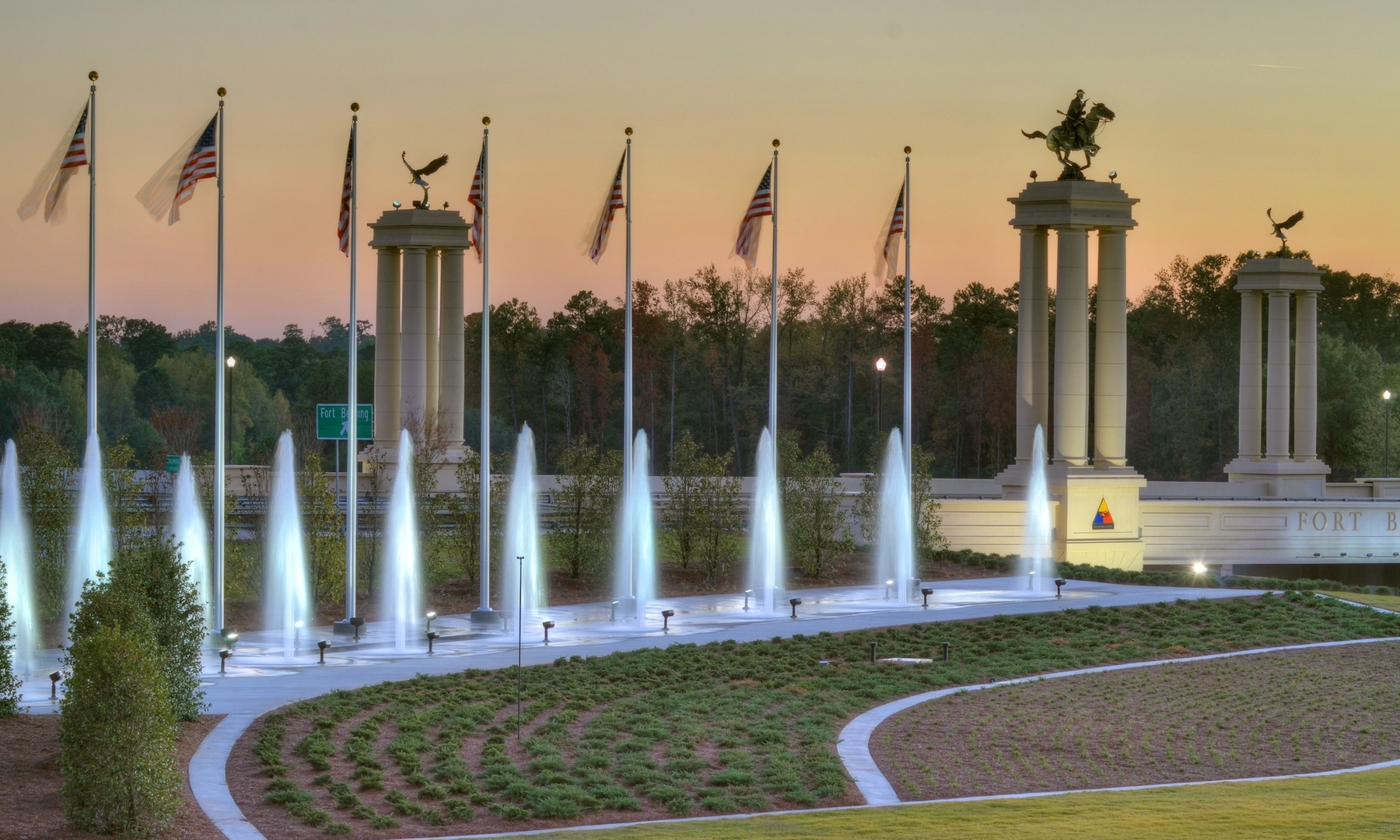 Fort Benning memorial entrance at sunset with American flags, fountains, and monument columns